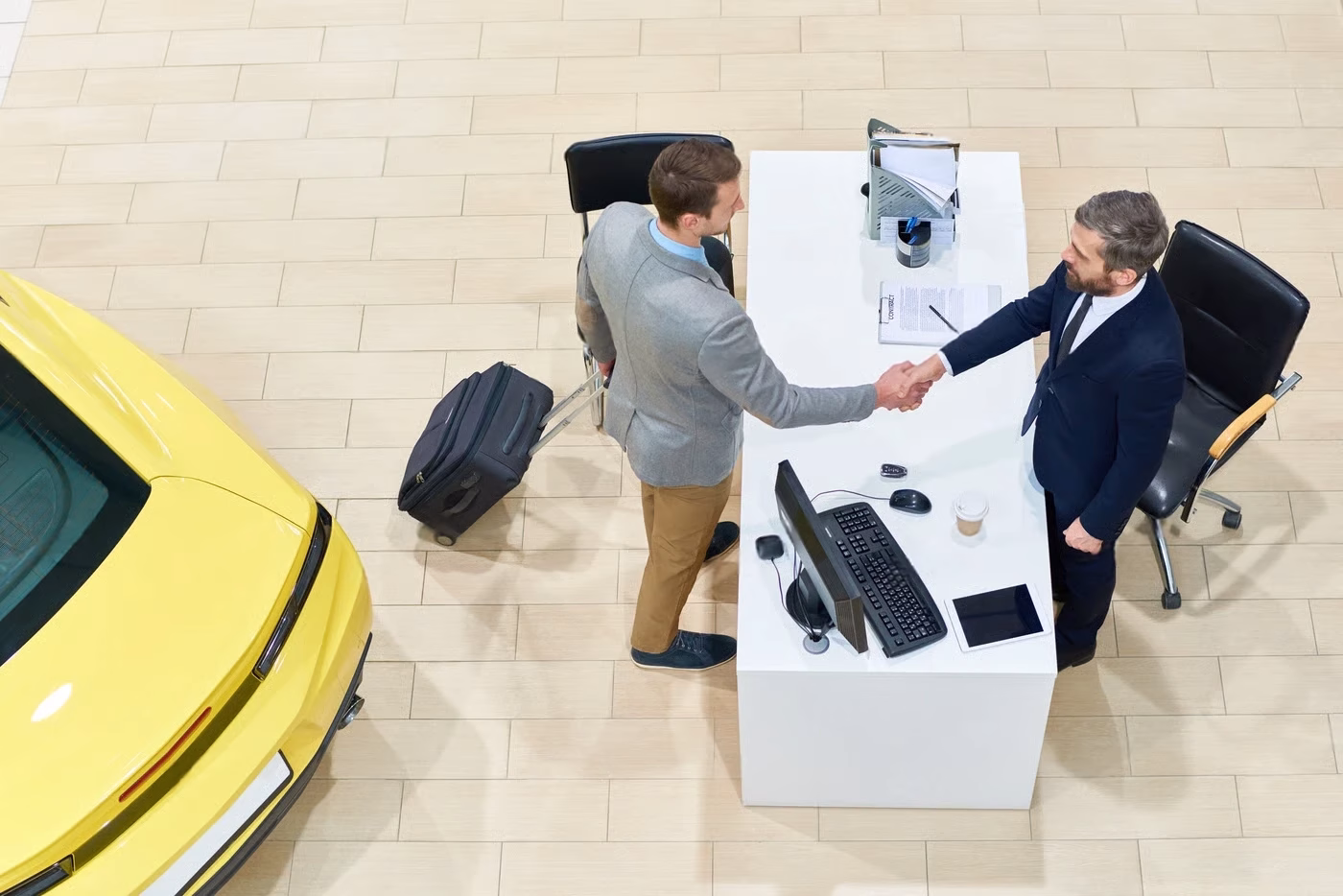 Two men in suits shake hands at a desk with a computer and contract, near a yellow car and luggage, in a modern office with tiled flooring.